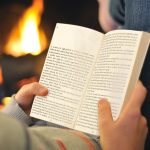 Woman in jeans and creen top reading a paperback copy of Razor in front of an open fire