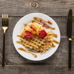 Plate with waffle, icecream and strawberries on wooden plank table and gold knife and fork
